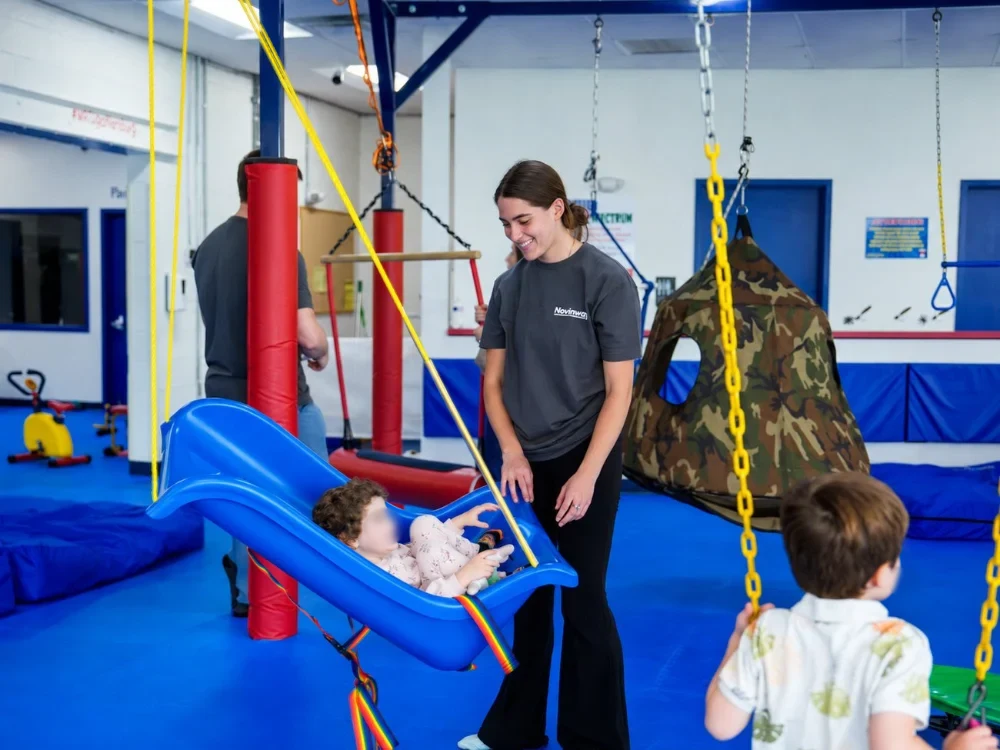 Naomi, a Novinway Care Specialist in a Novinway T-shirt, smiling beside a child on a sensory swing at a local sensory gym